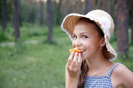 Portrait of preteen girl in straw hat eats apricot on green leaves backgroundの写真素材