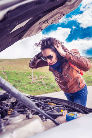 woman has a car breakdown on a mountain road and looks at the engine desperately waiting for roadside assistanceの写真素材