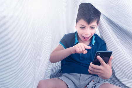 smartphone addiction of a child under white striped sheets holding a smartphone in his hand with very excited expressionの写真素材