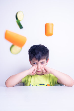 child who hates vegetables useful for his growth is sitting on the table with an angry look and the vegetables fly through the air on a white backgroundの写真素材