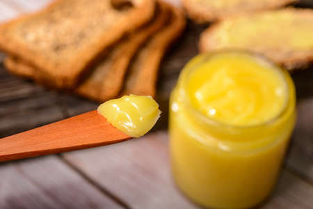 Ghee butter in glass jar with wooden spatula and sliced bread on wooden table.の写真素材