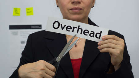 Female office worker or business woman cuts a piece of paper with the word overhead on it as an overhead reduction business concept.の写真素材