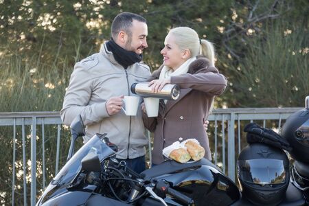 Portrait of pretty young couple drinking coffee and chatting near motorcycle