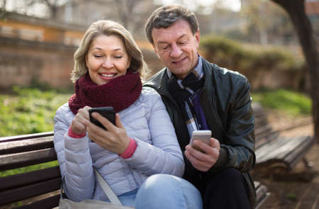 Happy mature couple with mobile phones on bench in park in autumnの写真素材