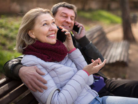 elderly couple talking on the phone on a park bench  in autumnの写真素材