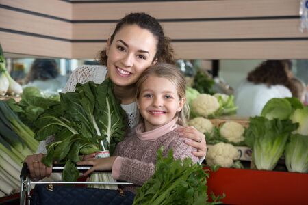portret of young smiling woman with beautiful blonde daughter choosing green vegetables at storeの写真素材