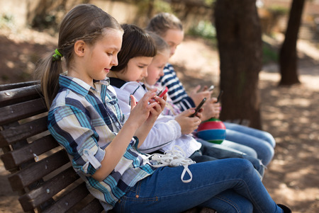 girls and boy with smartphones on bench in parkの写真素材