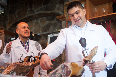 Mature butcher slicing jamon and smiling in delicatessen storeの写真素材