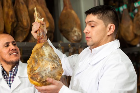 attentive technologist with butcher checking drying wurst and jamon at factoryの写真素材