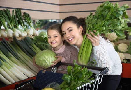 portret of  positive young woman with beautiful blonde daughter choosing green vegetables at storeの写真素材