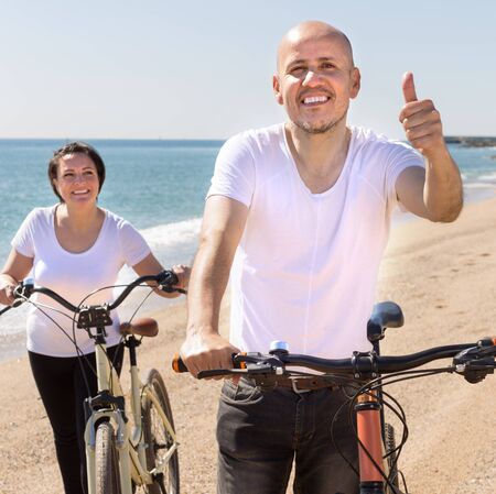 mature man and female walking with bicycles along the sea in summerの写真素材