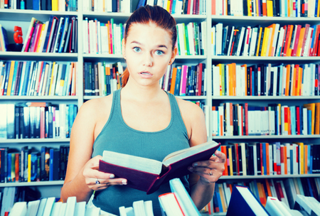 surprised teenager girl brunete looks for the right book in libraryの写真素材