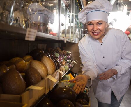 Mature female posing with fine chocolate eggs and cakes for saleの写真素材