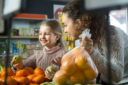 portret of smiling  young woman with beautiful blonde daughter choosing mandarins at storeの写真素材