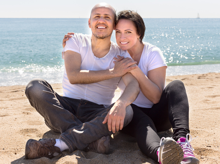 smiling elderly man with a woman in white shirts sitting on sand on beachの写真素材