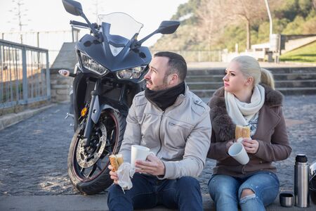 Pretty young couple drinking coffee and chatting near motorcycleの写真素材