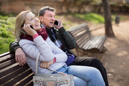 middle aged couple talking on the phone on a bench in the park in autumnの写真素材
