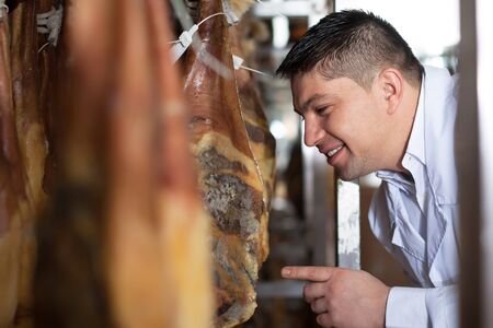 smiling butcher checking drying wurst and jamon at factoryの写真素材