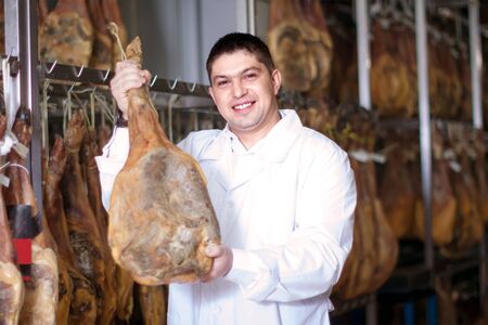 smiling mature butcher posing with jamon joints at meat factoryの写真素材