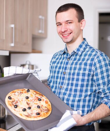 Young smiling man posing with delicious pizza in kitchen at homeの写真素材