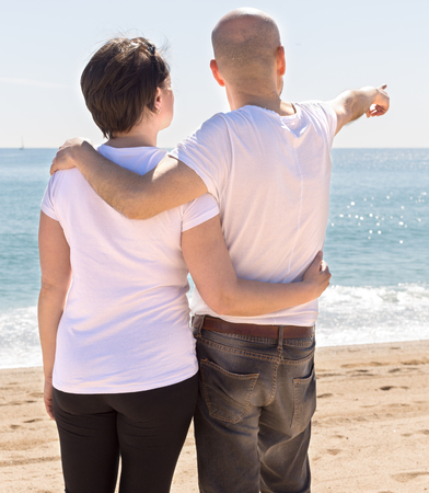 elderly man hugs a woman on the beach and indicates into the distance. view from the backの写真素材