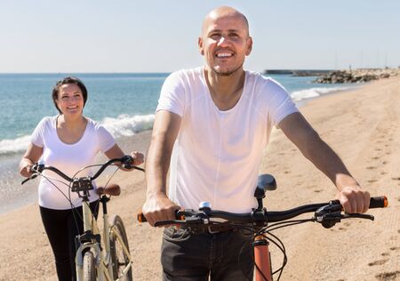 happy mature man and female walking with bicycles along the sea in summerの写真素材