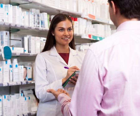 woman in a white robe salesman with a notebook in his hands in the pharmacy looking at visitorの写真素材