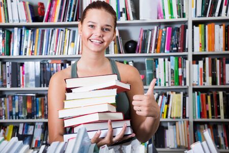 nice teenager girl holding stack of books shows thump up in a bookstoreの写真素材