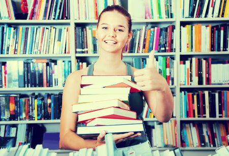 teenager girl holding stack of books shows thump up in a bookstore shows thump upの写真素材