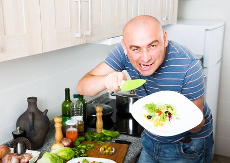 European emotional man with a knife and a plate of salad in his hand in the kitchenの写真素材