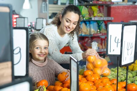smiling mother and blonde daughter buying mandarins in shop. focus on girlの写真素材