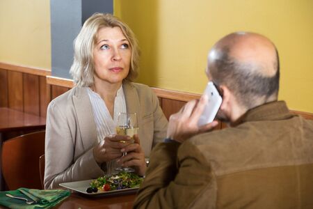 elderly blonde woman bored in a restaurant while her man talking on the phoneの写真素材