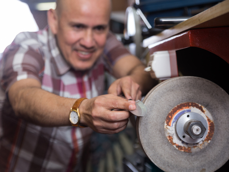Portrait of elderly specialist sharpening steel knife on  wheel lathe in workshop. Fokus on knifeの写真素材