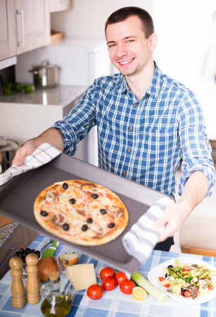 happy young man holding baking sheet with delicious pizza in kitchenの写真素材
