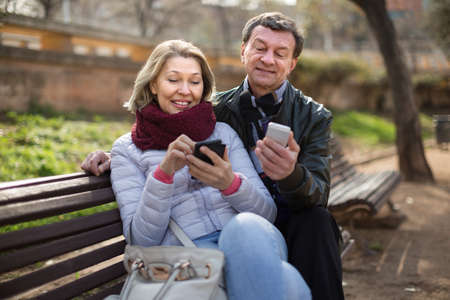 Portrait of Happy elderly couple with smartphones on park benchの写真素材
