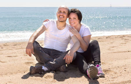 elderly man with a woman in white shirts sitting on sand on beach and smilingの写真素材