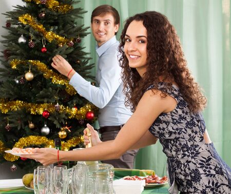 Young  happy couple decorating Christmas tree and serving  festive table in homeの写真素材
