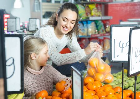 positive woman and little blonde girl posing whith mandarin in department fruits of store. focus on girlの写真素材