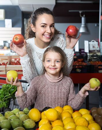 portret of positive young woman with beautiful daughter choosing apples at storeの写真素材