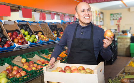 glad mature male vendor posing with apples in grocery storeの写真素材