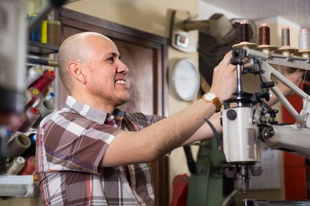 Mature Professional workman sewing leather boots on stitch lathe in workshopの写真素材