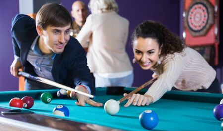 Group of friends playing billiards and smiling in night club. Focus on the billiard ball の写真素材