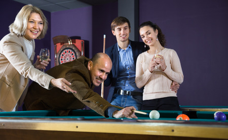men and women of different ages playing billiards and smiling in billiard room. focus on mature man