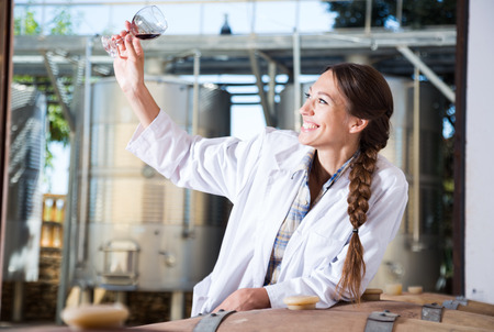girl specialist in white coat examines glass of wine on the background of barrels for fermentationの写真素材