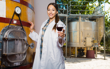 girl specialist in white coat examines glass of wine on the background of barrels for fermentationの写真素材