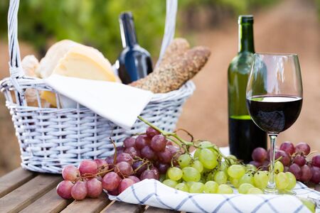 glass of red wine ripe grapes and picnic basket on table in vineyardの写真素材