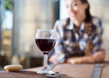 girl looking at glass of wine standing on wooden barrel in wineryの写真素材