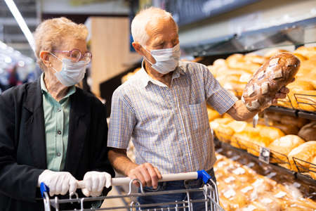 older european couple wearing mask and gloves with covid protection chooses buns and bread in...