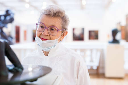 mature woman in mask protecting against covid examines paintings on display in hall of art museumの写真素材