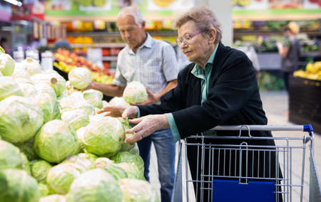 mature caucasian woman with glasses picks a head of cabbage in vegetable section of supermarketの写真素材